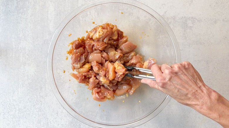 Combining chicken thigh chunks and marinade in glass bowl with tongs