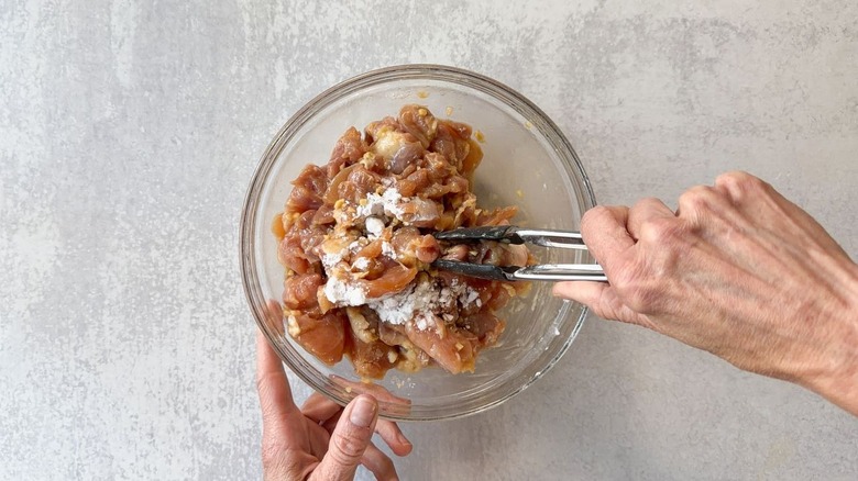 Tossing marinated chicken thigh chunks with cornstarch in glass bowl using tongs