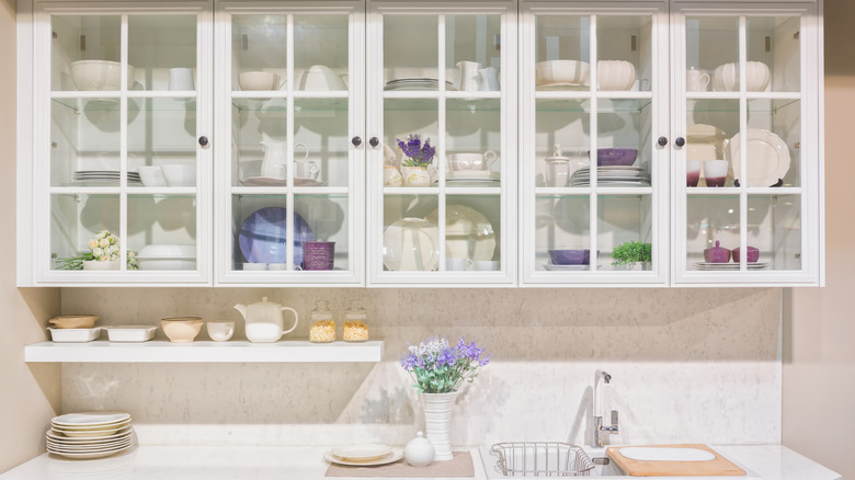 White kitchen cabinets with glass-front doors over a white counter with flowers in a vase