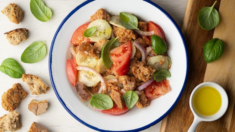 classic panzanella salad sitting in a white bowl on a counter surrounded by crusted bread pieces, basil, and olive oil