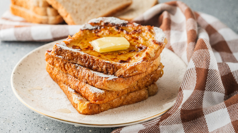 four pieces of french toast sitting on a white plate next to a kitchen towel. the toast is topped with butter and powdered sugar