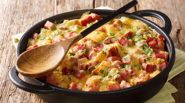 breakfast casserole with ham, cheese, and green onions in a skillet with a spoon laying on top