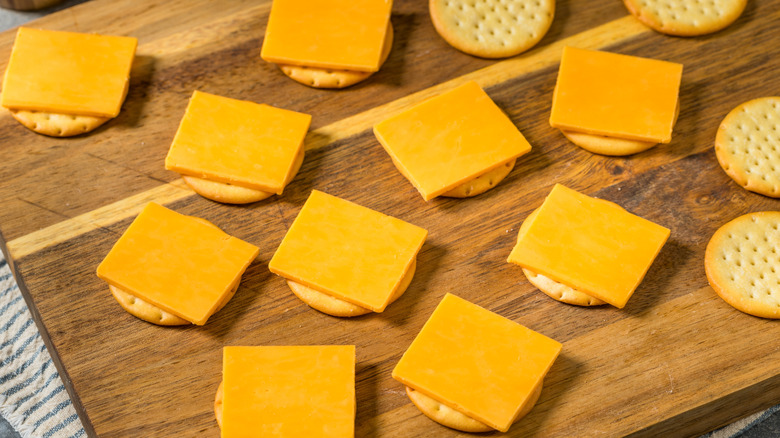 Various crackers topped with American cheese on wood cutting board.