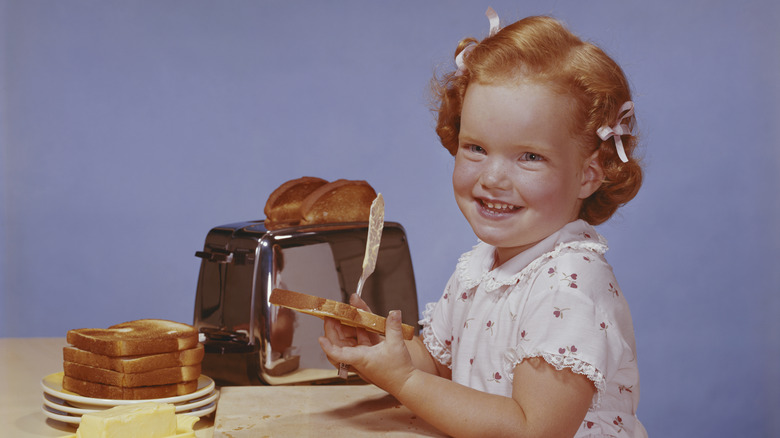 Little girl making peanut butter toast on table with a purple wall in background.