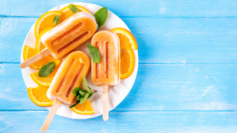 orange popsicles on a white plate with fresh orange and mint, presented ona blue wooden background.