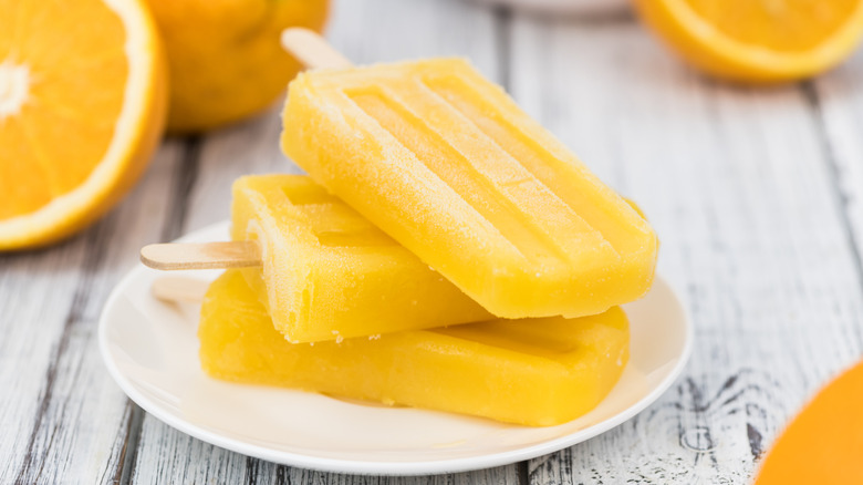 Orange popsicle on white plate with fresh orange in background on wooden table.