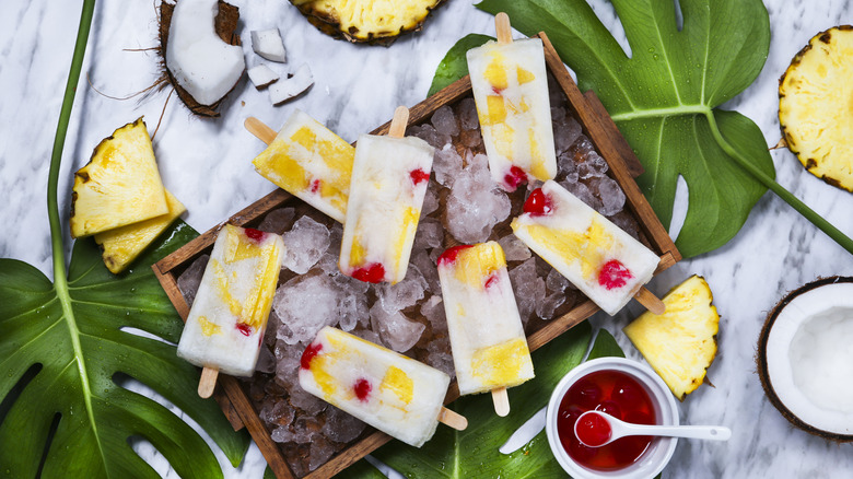 Pineapple coconut popsicles with cherries on a wooden tray of ice on a marble countertop surrounded by monstera leaves and fruit