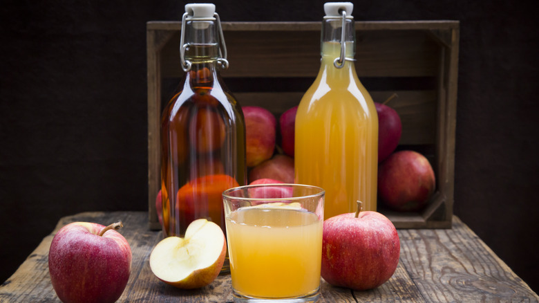 Atop a wooden table and with a dark background, a glass of apple cider is flanked by apples, both whole and sliced, and two glass bottles of apple cider, one hazy and one clear