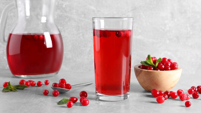 A glass of cranberry juice alongside a bowl of cranberries, a jug of juice, and several loose berries scattered across a grey surface