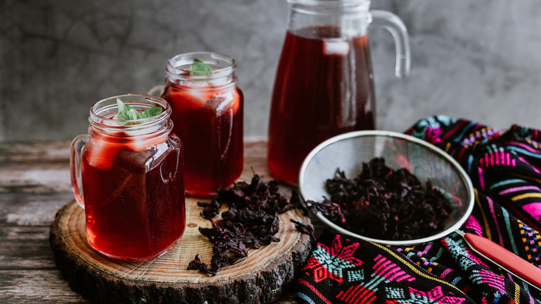 Mason jars and a jug filled with a hibiscus drink, as well as hibiscus flowers in a strainer, on a rustic wooden coaster with an embroidered Mexican cloth on the side