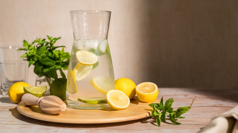 A carafe of lemonade on a wooden cutting board, surrounded by sliced lemons, mint leaves, and a wooden hand juicer
