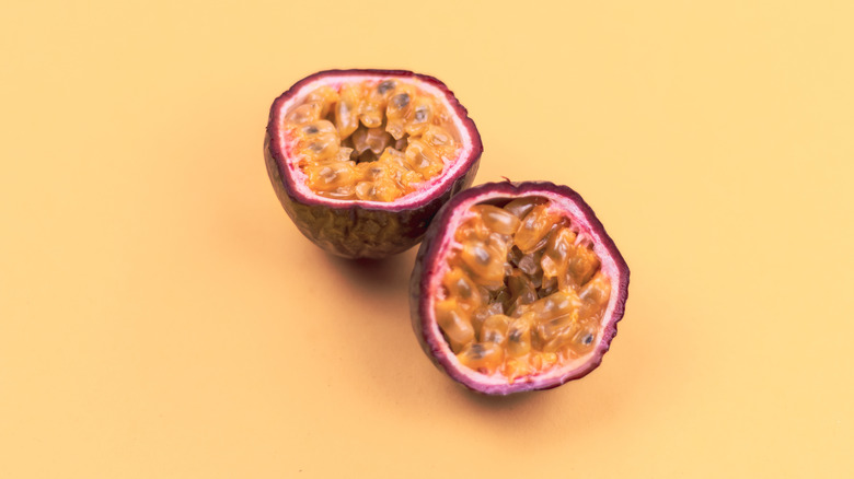 Two halves of a passionfruit rest on a peach-colored surface, showing the purple skin and yellowish pulp around the seeds