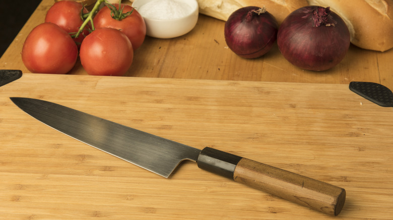 Close-up of a Japanese gyuto chef's knife on a cutting board with onions, bread, and tomatoes in the background