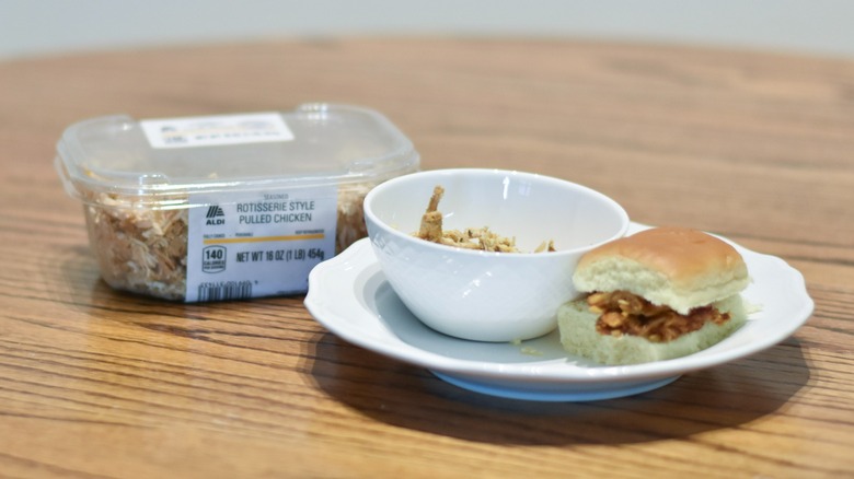 A container of rotisserie style pulled chicken next to a bowl of pulled chicken and a white plate with a barbecue pulled chicken sandwich