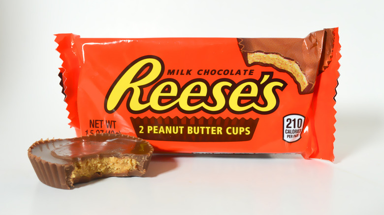A Reese's Peanut Butter Cup sits, bit into, next to its packaging