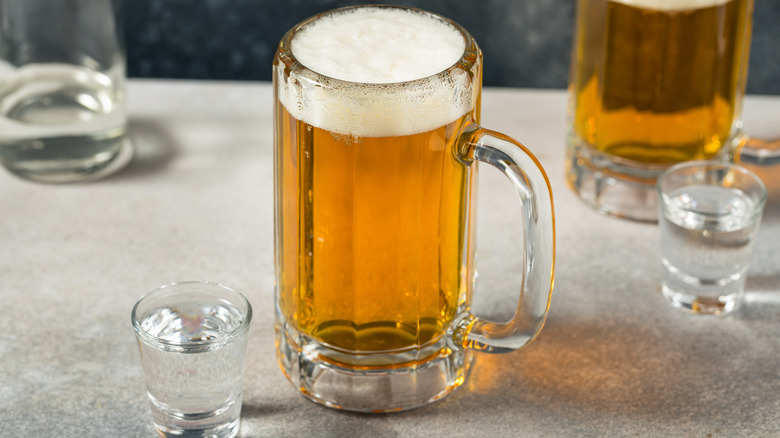 A frothy beer in a mug next to a clear shot in a shot glass on a table with other drinks in background
