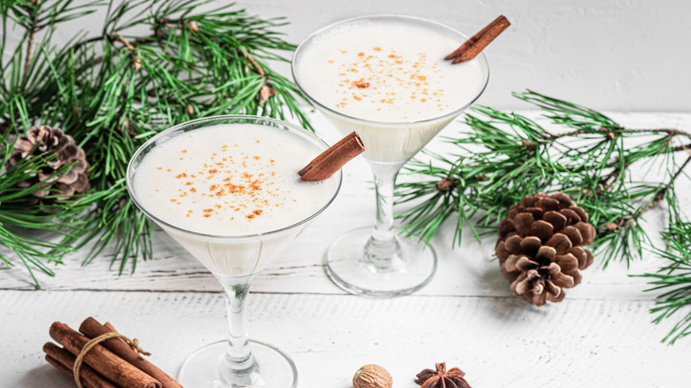 Two Brandy Alexander cocktails in glasses surrounded by pine branches, pinecones, a bundle of cinnamon sticks, and nutmeg