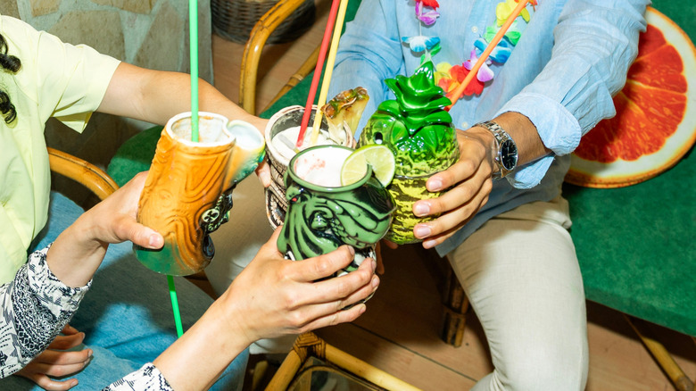 Three people sitting on wicker furniture and clinking tiki drinks together at a bar