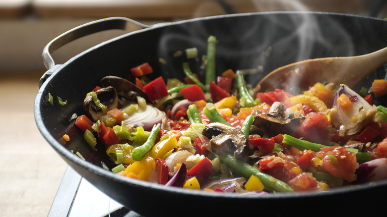 Vegetables cooking in a wok with steam rising and a wooden spoon being used to stir