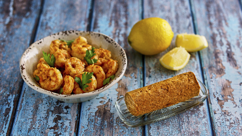 Bowl of sautéed shrimp with roll of red compound butter and lemons on the side
