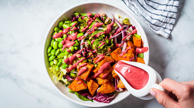 Bowl of salad with lima beans, lettuce, red onion, red cabbage and roasted sweet potatoes in a white bowl being drizzled with a creamy red dressing over marble background