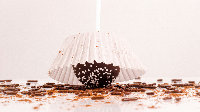 A chocolate cake pop with white nonpareil sprinkles in a cupcake liner upside down on a table of chocolate sprinkles.