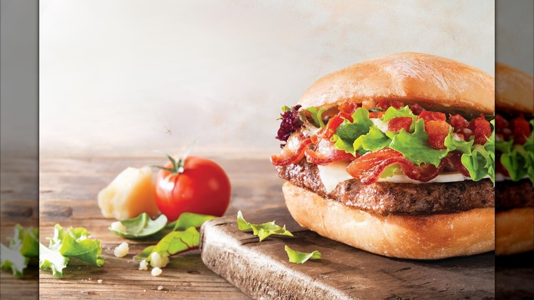 Wendy's Ciabatta Bacon Cheeseburger on a wooden cutting board. There's a ripe tomato, chunk of cheese, and leafy greens next to the cutting board.