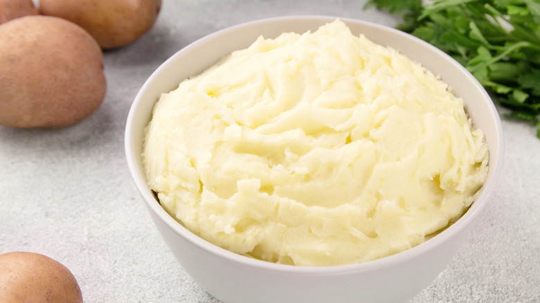 A bowl of mashed potatoes sits next to whole potatoes and fresh herbs.