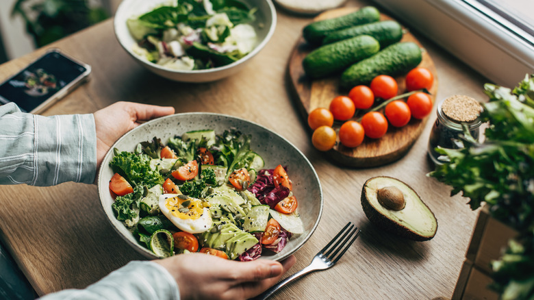 A person holding a bowl of vegetarian salad with more fresh salad ingredients in the background
