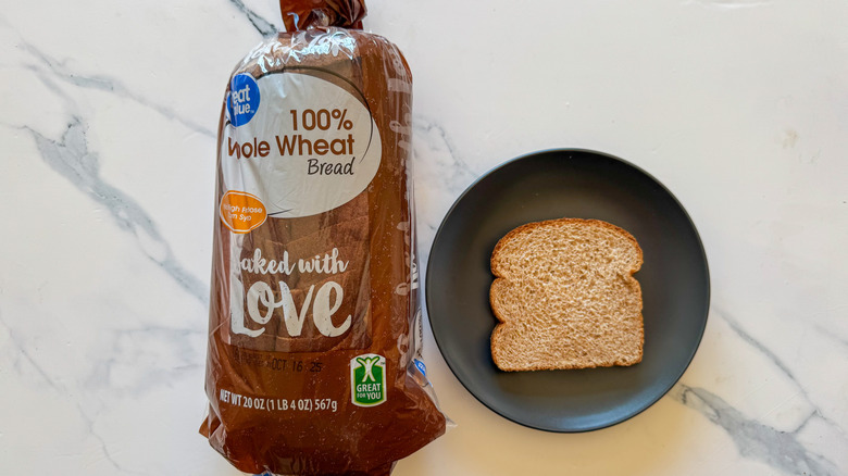 Package of Great Value whole wheat bread next to a slice of bread on a black plate on a marble countertop