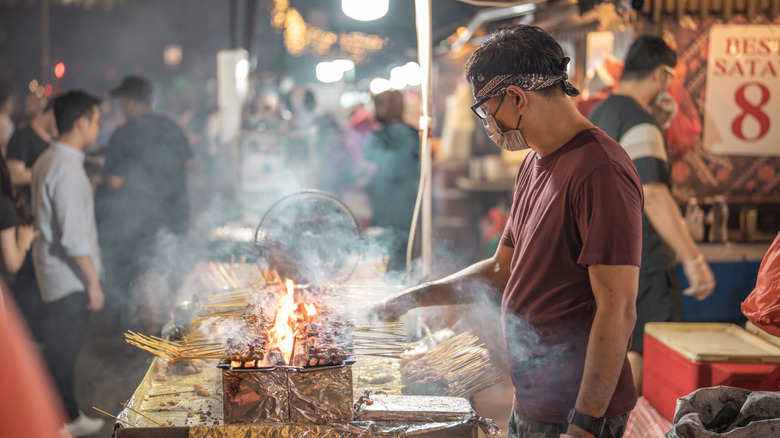 A vendor in Singapore grilling meat skewers on an open flame in a busy street market
