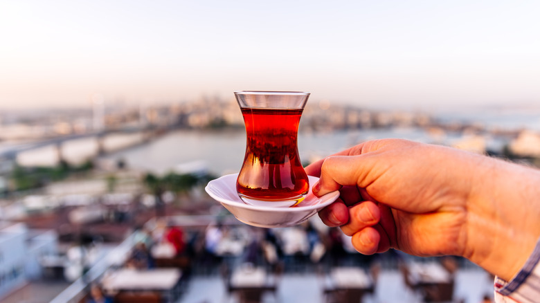 A hand holding a saucer atop which rests a glass of Turkish tea, with a blurred cityscape in the background