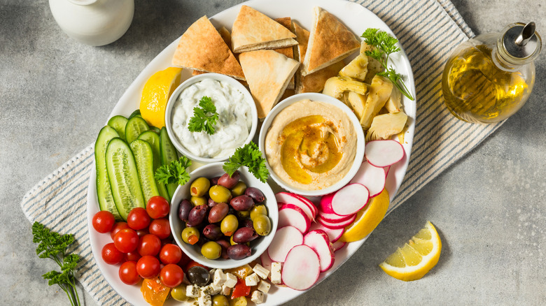 A mezze plate containing various types of vegetables, dips, olives, and flatbread, with lemons and olive oil on the side