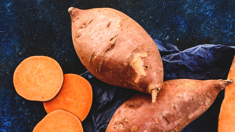 Bright orange sweet potatoes, some peeled and sliced into discs and some whole and unpeeled, on a dark blue background.
