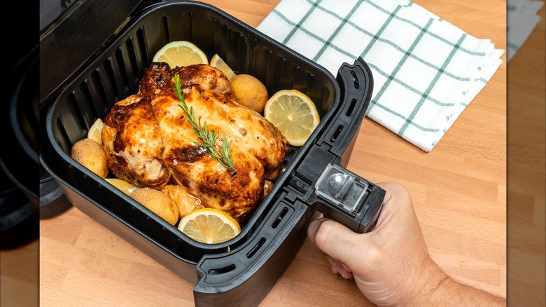An air fryer cooking basket containing a roast chicken, potatoes, lemon and a sprig of rosemary. In the background there is a checkered dish towel.