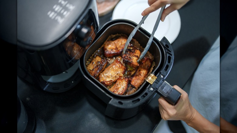 A top down view of an air fryer cooking basket containing pork ribs. A person is hold the cooking basket and turning the ribs with silicone tongs.
