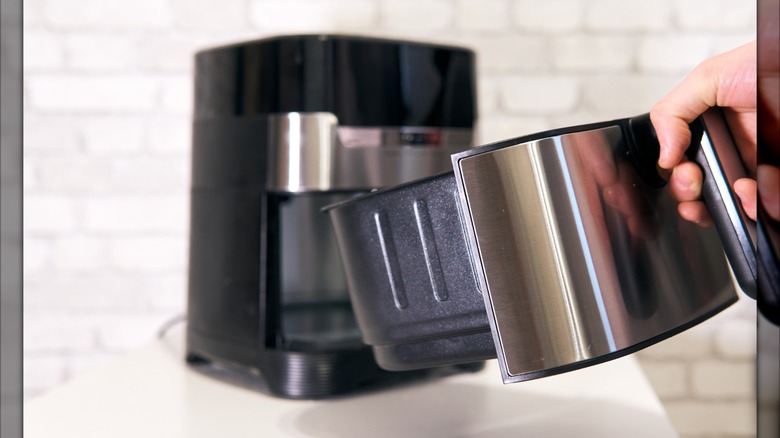 An air fryer basket is being held with a black air fryer in the background.