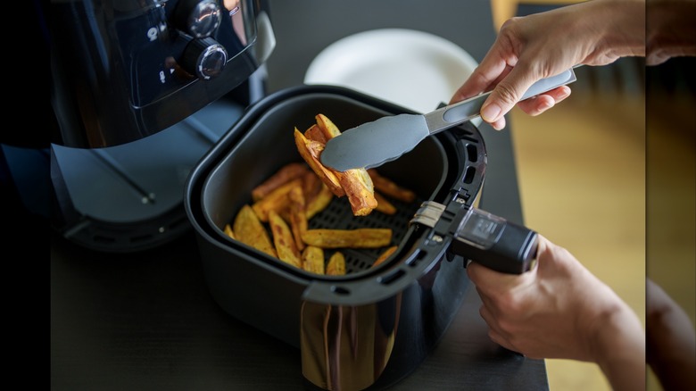 A person hold several roasted potato chips with a pair of silicone tongs. The air fryer basket contains more roasted potatoes.