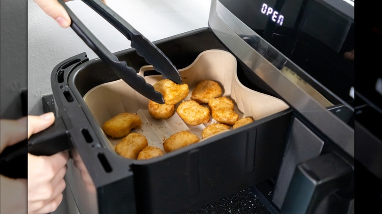 An air fryer cooking basket containing chicken nugget. One nugget is being held by a pair of black silicone cooking tongs.