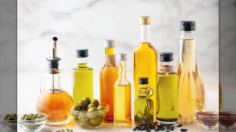 A counter top has eight bottles of oil on it. There is also a bowl of olives and other bowls with different seeds and spices.