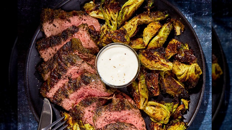 A black plate with sliced seared steak, brussel sprouts, and a small dish of horseradish cream