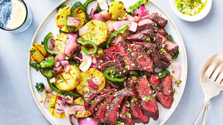 A white plate with grilled steak, potato and pepper salad, and herb dressing