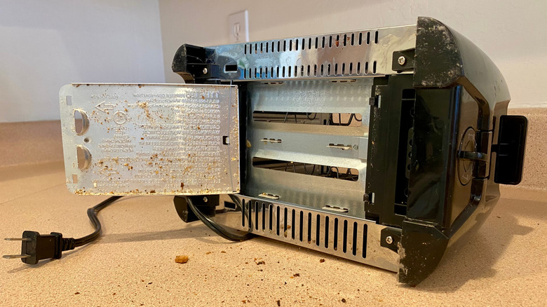 A chrome and black plastic toaster lying on its side, bottom visible, crumb tray open, with crumbs spilled on the counter