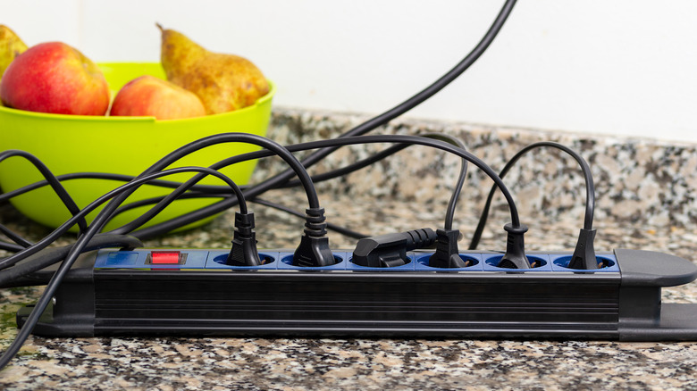 An extension cord and power strip with several cords plugged in, in front of a green bowl holding fruit