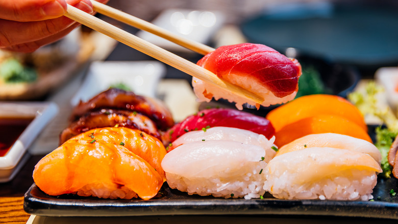 Pieces of sushi on a black plate with one being picked up with a pair of chopsticks