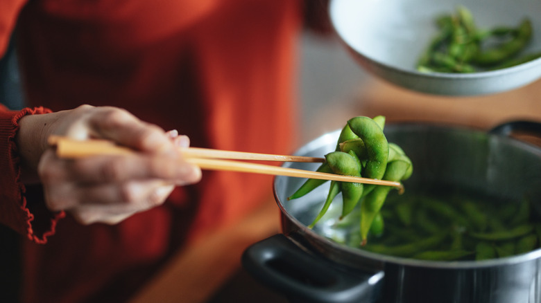 A person holding edamame from a pot on the stove with chopsticks