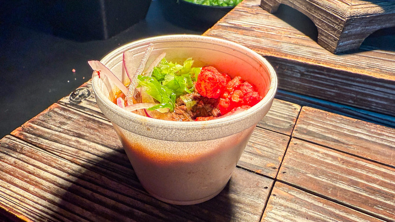 A white styrofoam cup of birria ramen garnished with onions and Flamin' Hot Cheetos