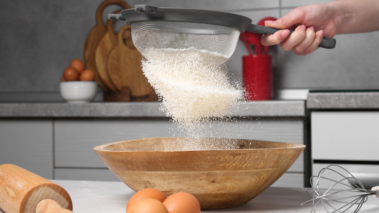 Person sifting flour into a wooden bowl