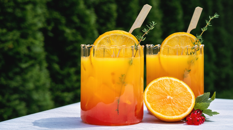 Orange juice and mixers in two glasses with thyme sprig garnishes and half an orange and red berries beside them