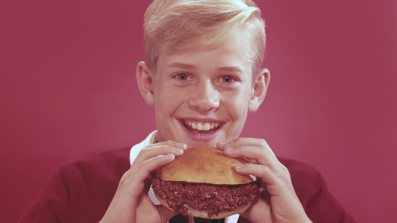 A boy smiling and holding a Sloppy Joe against a red background